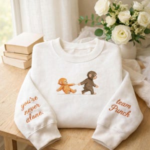 May include: White sweatshirt featuring an embroidered illustration of two monkeys, one orange and one brown, holding hands. The left sleeve reads "you're never alone," and the right sleeve says "team Punch" in orange thread.