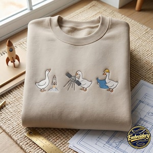 Puede incluir: Una sudadera de cuello redondo en color beige con un bordado de tres ocas blancas. La primera oca observa el lanzamiento de un cohete, la segunda maneja un satélite y la tercera lleva un casco de obra amarillo mientras sostiene un plano azul.