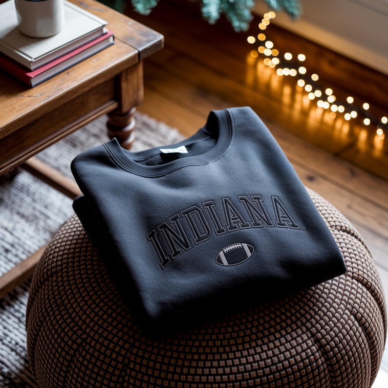 K&ouml;nnte beinhalten: Ein dunkelgraues Sweatshirt mit dem Wort "INDIANA" in einem Bogen &uuml;ber der Brust in dunklerem Grau und einer Football-Grafik. Das Sweatshirt liegt gefaltet auf einem braunen, gewebten Hocker.