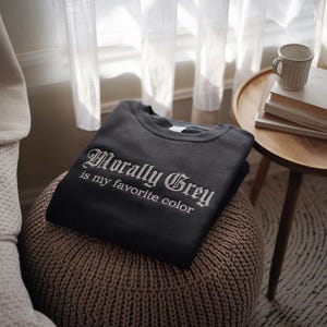 以下が含まれることがあります： 白いゴシック体で「Morally Grey is my favorite color」と書かれた、濃いグレーのスウェットシャツ。スウェットシャツは折りたたまれ、織りのオットマンの上に置かれています。マグカップと本が置かれた小さなサイドテーブルが背景にあります。
