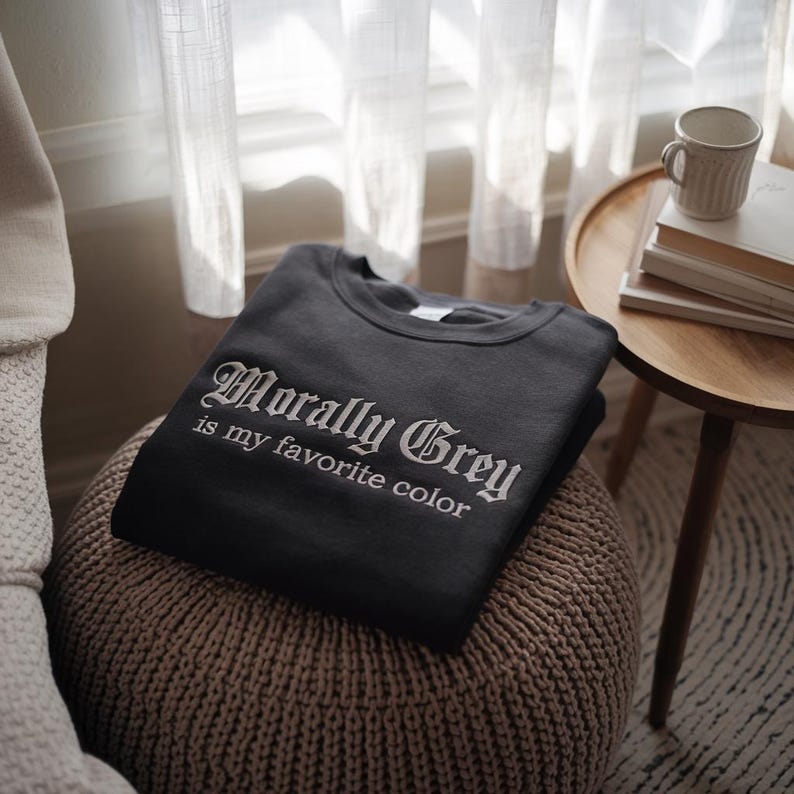 以下が含まれることがあります： 白いゴシック体で「Morally Grey is my favorite color」と書かれた、濃いグレーのスウェットシャツ。スウェットシャツは折りたたまれ、織りのオットマンの上に置かれています。マグカップと本が置かれた小さなサイドテーブルが背景にあります。