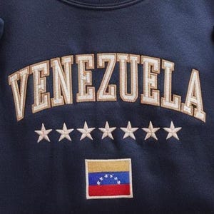Op de afbeelding: Marineblauw sweatshirt met het woord "VENEZUELA" geborduurd in gouden en witte letters in collegestijl. Onder de tekst staan zeven geborduurde sterren en een kleine Venezolaanse vlagpatch.