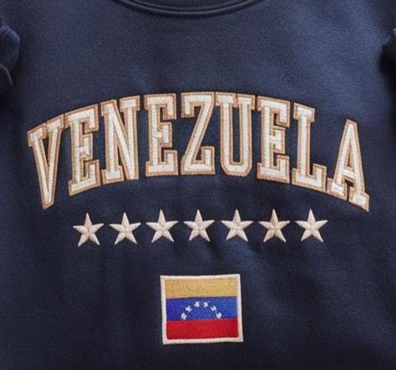 Op de afbeelding: Marineblauw sweatshirt met het woord "VENEZUELA" geborduurd in gouden en witte letters in collegestijl. Onder de tekst staan zeven geborduurde sterren en een kleine Venezolaanse vlagpatch.