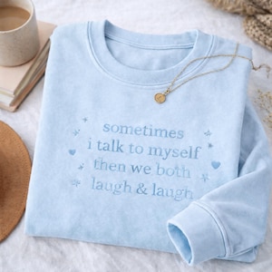 Puede incluir: Sudadera azul claro con la frase bordada "sometimes I talk to myself then we both laugh & laugh". Un collar dorado con un pequeño colgante descansa sobre la sudadera doblada.
