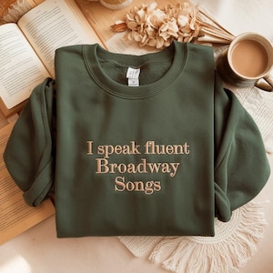May include: An olive green sweatshirt with the embroidered text "I speak fluent Broadway Songs." The crew neck sweatshirt is folded on a light-coloured surface, with a book and a cup of coffee in the background.