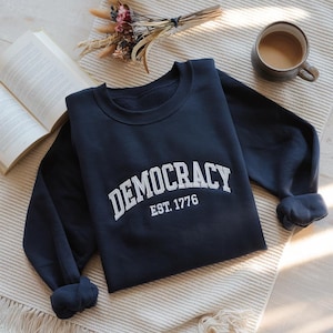 Op de afbeelding: Een marineblauw sweatshirt met witte geborduurde tekst die "DEMOCRACY EST. 1776" luidt.