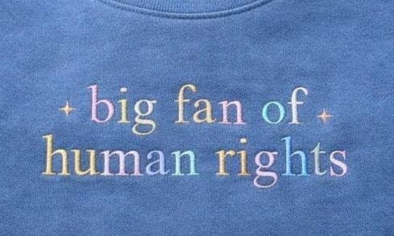 May include: Close-up of a blue sweatshirt with the embroidered text "big fan of human rights" in pastel colors. The text is centered and the sweatshirt has a crew neck.