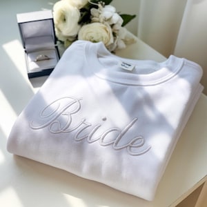 May include: White sweatshirt with the word "Bride" embroidered in white cursive lettering.