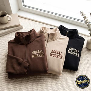 Puede incluir: Tres sudaderas con cremallera de un cuarto en marrón oscuro, beige y negro. Cada sudadera presenta el texto SOCIAL WORKER bordado en el pecho izquierdo. Las prendas tienen un cuello alto con cierre de cremallera metálica y mangas largas.