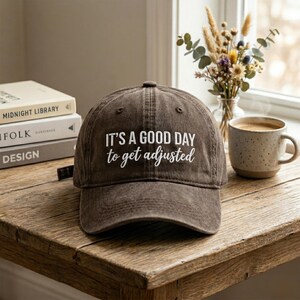 Peut inclure: Casquette de baseball marron avec le texte "IT'S A GOOD DAY to get adjusted" en blanc. Le chapeau est posé sur une table en bois, à côté de livres et d'une tasse de café. Accessoire décontracté.