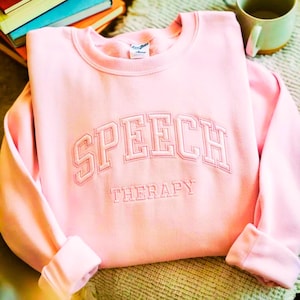 May include: A light pink crewneck sweatshirt featuring the embroidered text SPEECH THERAPY in a collegiate font. The word SPEECH is arched in large block letters, while THERAPY is written in smaller, straight block letters below it.