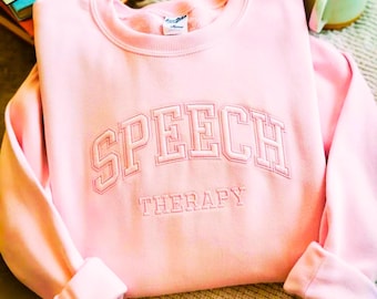 Può includere: Una felpa girocollo rosa chiaro con il testo ricamato SPEECH THERAPY in un carattere in stile college. La parola SPEECH è arcuata in grandi lettere stampatello, mentre THERAPY è scritta in lettere stampatello più piccole e dritte.