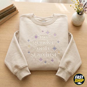 May include: A beige crewneck sweatshirt featuring embroidered text that reads no gender only stardust in a serif font. The design is surrounded by small, multi-colored embroidered star shapes in shades of purple, blue, and yellow.