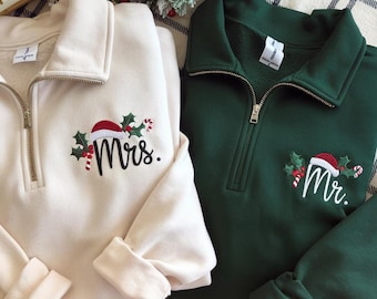 May include: Two quarter-zip sweatshirts, one cream and one dark green. The cream sweatshirt has "Mrs." embroidered with a Santa hat and holly. The green sweatshirt has "Mr." embroidered with a Santa hat and holly. Both have gold zippers.