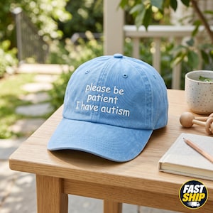May include: A light blue baseball cap with the text "please be patient I have autism" embroidered in white. The cap is sitting on a wooden table. A small notebook and pencil are also on the table.