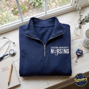 May include: A navy blue quarter-zip sweatshirt featuring white embroidered text that reads CUSTOM UNIVERSITY NURSING, with the letter U replaced by a stethoscope icon and the letter I replaced by a syringe icon on the left chest area.