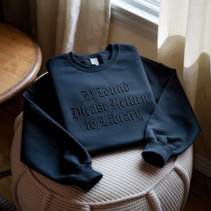 May include: A navy blue sweatshirt with the embroidered text "If Found Please Return to Library" in a gothic font. The sweatshirt is folded on a beige patterned ottoman, with a wooden table and window in the background.