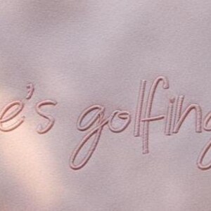 May include: A close-up of a light pink fabric with the embroidered phrase "he's golfing" in a matching pink thread. The text is in a cursive font. The background has soft shadows.