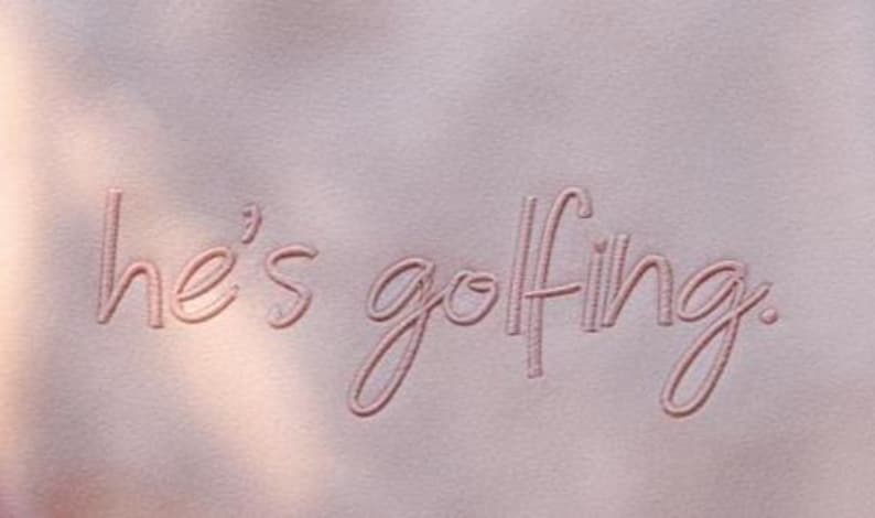 May include: A close-up of a light pink fabric with the embroidered phrase "he's golfing" in a matching pink thread. The text is in a cursive font. The background has soft shadows.