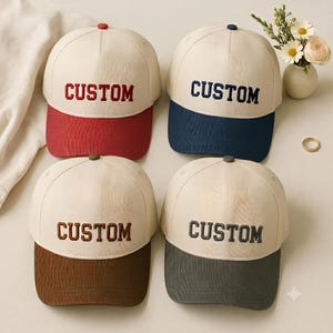 May include: Four baseball caps with off-white crowns and coloured brims. Each cap features the word CUSTOM embroidered in a matching colour on the front panel. The brims and embroidery are red, navy blue, brown, and dark grey respectively.