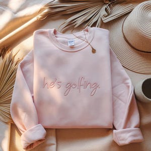 May include: A light pink sweatshirt with the embroidered phrase "he's golfing" in a cursive font. The long-sleeved crewneck sweatshirt is folded, with the cuffs turned up. A gold necklace and a straw hat are visible in the background.