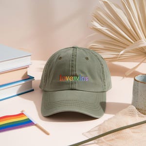 May include: An olive green baseball cap with the embroidered text "lovewins" in rainbow colors. The cap is displayed on a light pink surface with a small rainbow flag and books in the background.