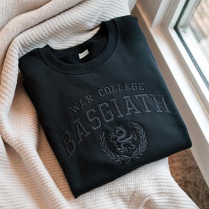 May include: Black sweatshirt featuring the embroidered text "WAR COLLEGE BASGIATH" in grey. A crest with a dragon and laurel wreath is below the text. The sweatshirt is folded on a cream-coloured blanket.