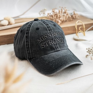 Puede incluir: Gorra de béisbol gris oscuro con el texto bordado "MINNESOTA STRONG!". La gorra tiene una visera curva y un botón en la parte superior. El sombrero está hecho de una tela lavada y desgastada.