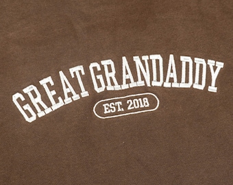 May include: Brown sweatshirt with the text "GREAT GRANDADDY" in white, arched across the front. Below the text is "EST. 2018" in a white oval. The sweatshirt is made of a soft material.