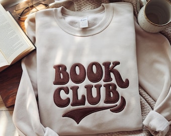 Puede incluir: Una sudadera beige con las palabras "BOOK CLUB" bordadas en marrón. La sudadera está doblada sobre una superficie de madera junto a un libro abierto, unas gafas y una taza.