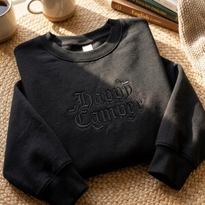 Peut inclure: Un sweat-shirt noir à col rond avec une broderie noire sur la poitrine indiquant Happy Camper dans une police gothique. Le motif comprend de petites chauves-souris brodées et des détails de toile d'araignée autour du texte, avec des manches longues.