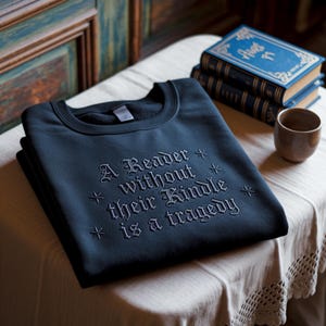 Może przedstawiać: Ciemnoniebieska bluza z haftowanym napisem "A Reader without their Kindle is a tragedy". Bluza jest złożona na stole z książkami i małym brązowym kubkiem.