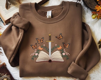 Op de afbeelding: Een bruine sweatshirt met een geborduurd ontwerp met een open boek, een zwaard, vlinders en bloemaccenten in oranje, groen en wit. De sweatshirt wordt gepresenteerd op een witte, zachte ondergrond.