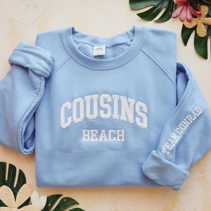 Peut inclure: Un sweat-shirt bleu clair avec les mots "COUSINS BEACH" en lettres blanches sur la poitrine. La manche porte l'inscription "TEAM CONRAD" en blanc. Le sweat-shirt est fait d'une matière douce.