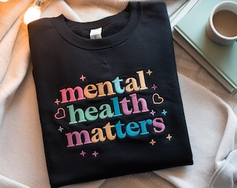 May include: Black sweatshirt with the embroidered text "mental health matters" in pink, blue, orange, and green. The sweatshirt is folded on a light-colored blanket, with a book and a mug of coffee in the background.