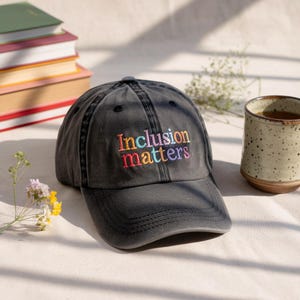 May include: A dark grey baseball cap with the embroidered text "Inclusion matters" in rainbow colours. The hat is on a white surface next to a cup of tea and a stack of books.