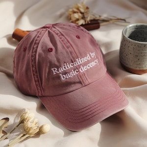 Op de afbeelding: Een stoffige roze baseballcap met de geborduurde witte tekst "Radicalized by basic decency". De cap heeft een gebogen klep en een zes-panelen ontwerp. De hoed is gemaakt van een zacht, versleten materiaal.