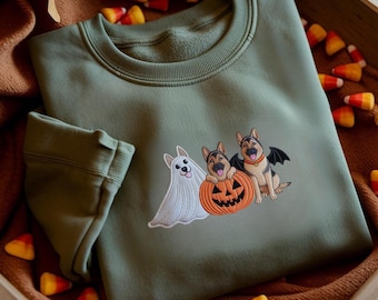 May include: An olive green crewneck sweatshirt featuring an embroidered graphic of three German Shepherd dogs. One dog is dressed as a ghost, one sits inside a carved orange pumpkin, and the third wears black bat wings for a Halloween theme.