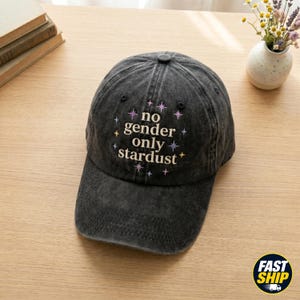 Könnte beinhalten: Eine anthrazitfarbene Baseballkappe mit verwaschener Optik, die den gestickten Schriftzug 'no gender only stardust' in Cremefarben zeigt, umgeben von kleinen, farbigen Sternsymbolen in Violett, Gelb und Hellblau auf der Vorderseite.