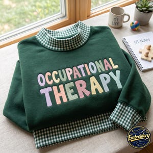 May include: A dark green sweatshirt featuring a mock neck collar and sleeve cuffs in a green and white gingham pattern. The front displays the text OCCUPATIONAL THERAPY embroidered in a multi-colored, pastel-toned, rounded, bubbly font style.