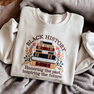 May include: A cream-coloured sweatshirt with an embroidered design of a stack of books with the words "Black History" above the books and "Honoring the past, inspiring the future" below the books.