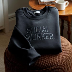 Könnte beinhalten: Ein dunkelgraues Sweatshirt mit dem gestickten Text "SOCIAL WORKER." Das Sweatshirt ist ordentlich auf einem braunen Hocker gefaltet. Der Ärmel ist teilweise sichtbar, und eine weiße Tasse steht auf einem kleinen Holztisch im Hintergrund.