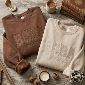 Pode incluir: Duas camisolas de gola redonda, uma castanha e uma creme, com o texto bordado BCBA EST. 2026 no peito. A camisola castanha tem o nome Kelsey bordado na manga, enquanto a camisola creme tem o nome Liam.