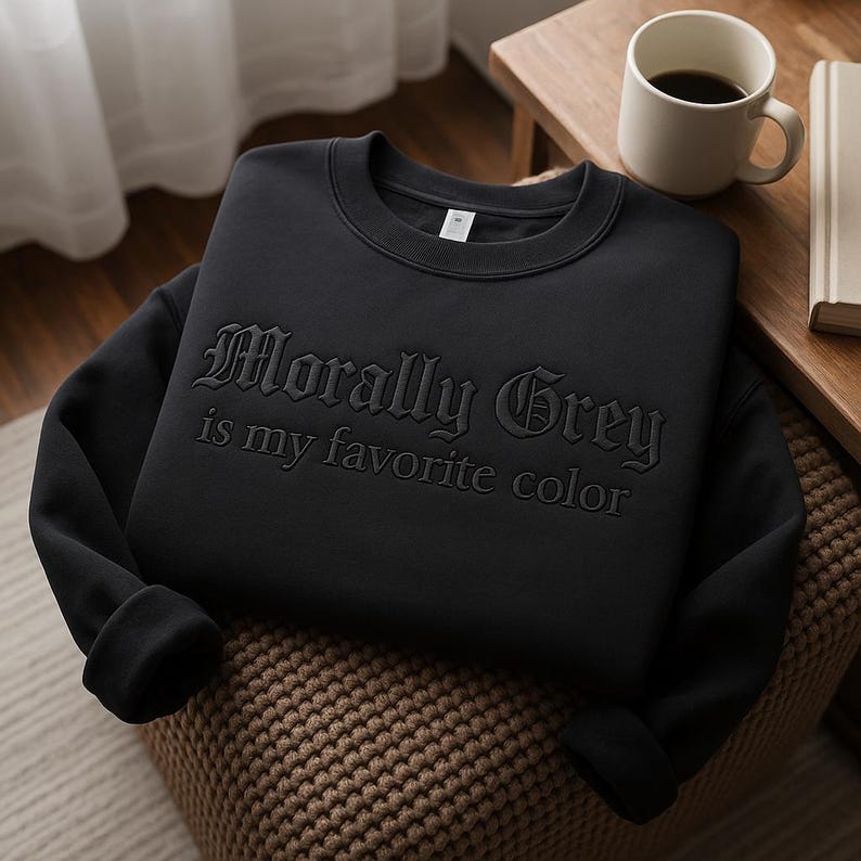 K&ouml;nnte beinhalten: Ein schwarzes Sweatshirt mit dem gepr&auml;gten Schriftzug "Morally Grey is my favorite color". Das Kleidungsst&uuml;ck liegt auf einer gewebten Oberfl&auml;che. Eine Kaffeetasse und ein Buch sind im Hintergrund zu sehen.