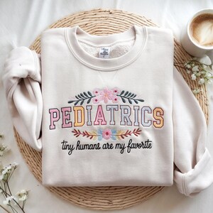 May include: A cream-colored sweatshirt with the words "PEDIATRICS" embroidered in colorful letters. The text "tiny humans are my favorite" is embroidered below the word "PEDIATRICS".