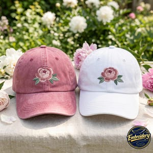 May include: Two baseball caps, one in a dusty rose color and one in white, each featuring an embroidered pink peony flower with green leaves centered on the front panel. Both caps have a curved brim and a structured six-panel design.