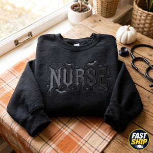 May include: A black long-sleeve sweatshirt featuring the word NURSE embroidered in a gothic font. The design includes embroidered spiderwebs and small bats surrounding the text. The sweatshirt has a crew neckline and ribbed cuffs.