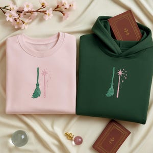 Op de afbeelding: Een lichtroze sweatshirt en een donkergroene hoodie, beide met geborduurde ontwerpen van een bezem en een toverstaf. De kledingstukken worden gepresenteerd op een cr&egrave;mekleurig oppervlak met decoratieve elementen.