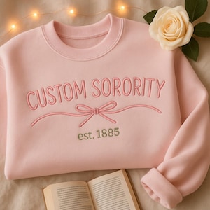 Op de afbeelding: Een lichtroze sweatshirt met de woorden "CUSTOM SORORITY" geborduurd in roze draad. Onder de tekst staat een strikdesign en het jaar "est. 1885" in groen. Een crèmekleurige roos en een open boek op de achtergrond.
