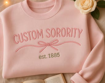 May include: A light pink sweatshirt with the words "CUSTOM SORORITY" embroidered in a matching pink thread. Below the text is a bow design, and the year "est. 1885" is embroidered in a muted green. A cream-colored rose and a book are in the background.